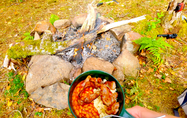 Frukost i skogen En hand som håller en skål med scrambled eggs, vitabörnor och bacon framför en öppen eld och stenar.