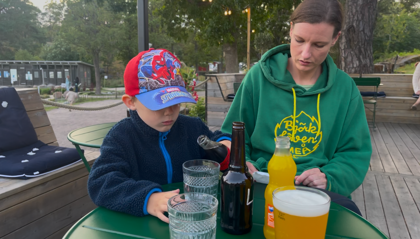 Algots på Udden En pojke och en kvinna sitter vid ett bord med drycker i en utomhusmiljö.