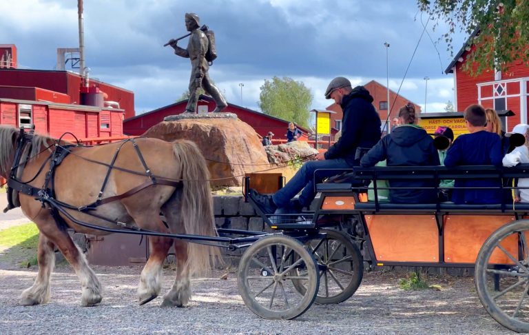 En hästdragen vagn med människor passerar en staty i en färgglad by.