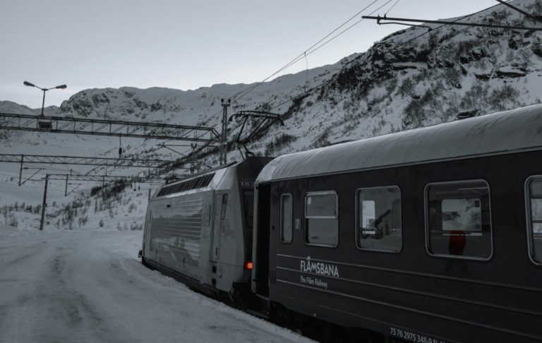 Tåg i snölandskap med berg i bakgrunden och låg sol.