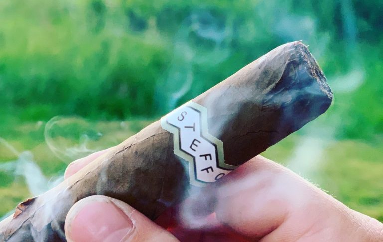 En hand håller en cigarr som ryker, med grön bakgrund.