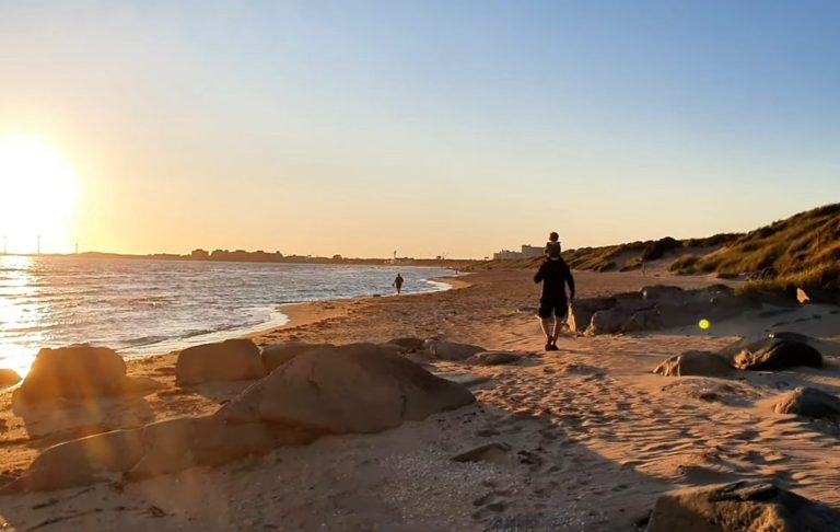 Person går längs en strand vid solnedgången, med vatten och klippor i bakgrunden.