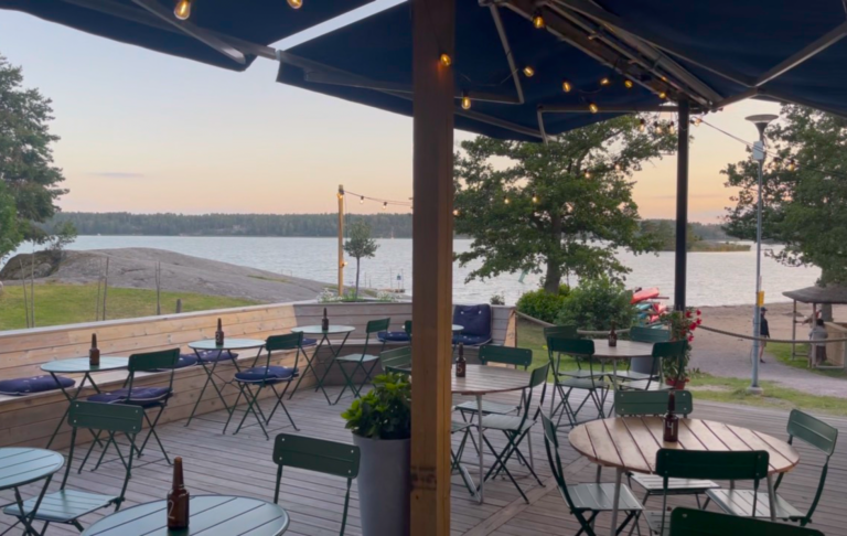 Uteplats vid vattnet med bord och stolar, omgiven av träd och en solnedgång.