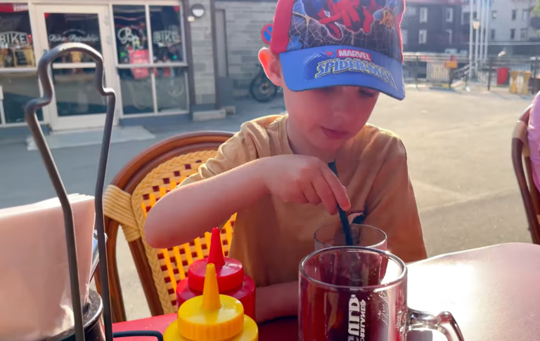 Pojke med blå hatt rör om i en kopp på en uteservering. Ketchup och senap syns i bakgrunden.