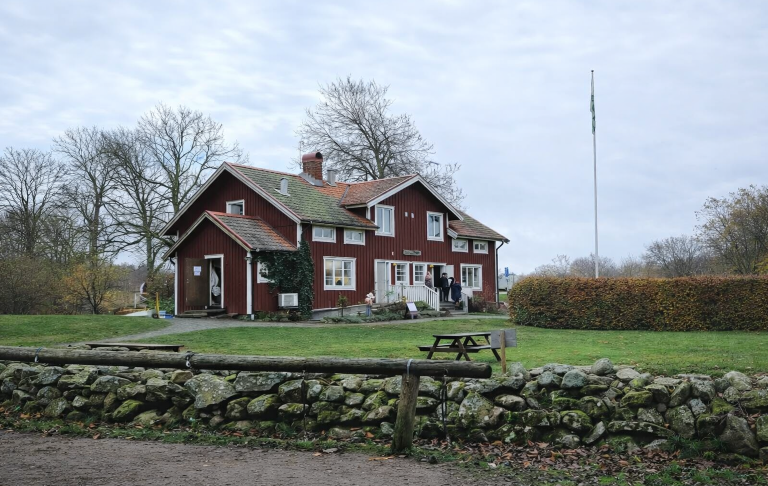 Röd stuga med grön tak, omgiven av träd och en marksten.