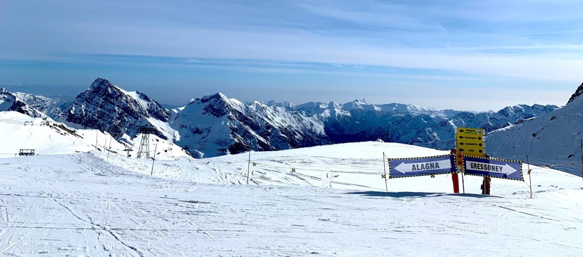 Vinterlandskap med snöklädda berg och en skidbacke i förgrunden. skidskyltar med pilar och texten Alagna & Gressoney.