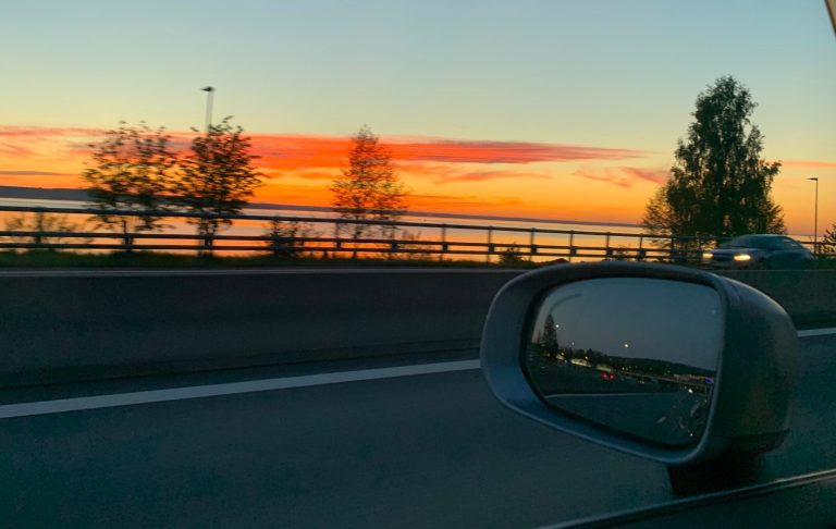 Solnedgång vid vägen, med träd och reflektioner i backspegelen.