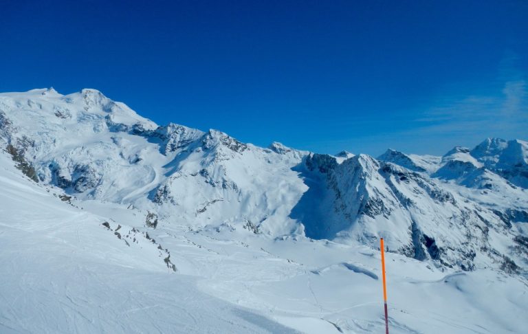 Snöklädda alptoppar med klarblå himmel och en färgglad pistpinne i förgrunden. Skidbacke.