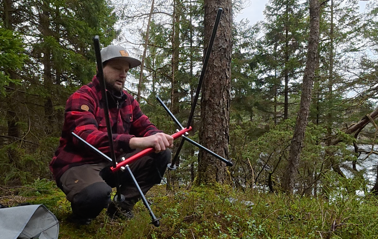 Person i röd- och svartrandig jacka som sitter på mossig mark vid en sjö och håller upp en ram av en campingstol i handen.
