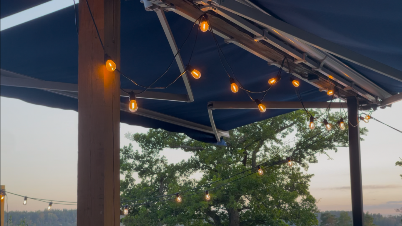 Algots på Udden Ljusande hängande lampor på en pergola med en trästolpe och träd i bakgrunden.