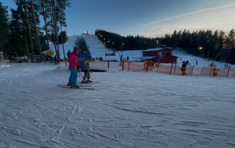 Två personer åker skidor på en snöig backe vid solnedgång. Skidområde i bakgrunden.