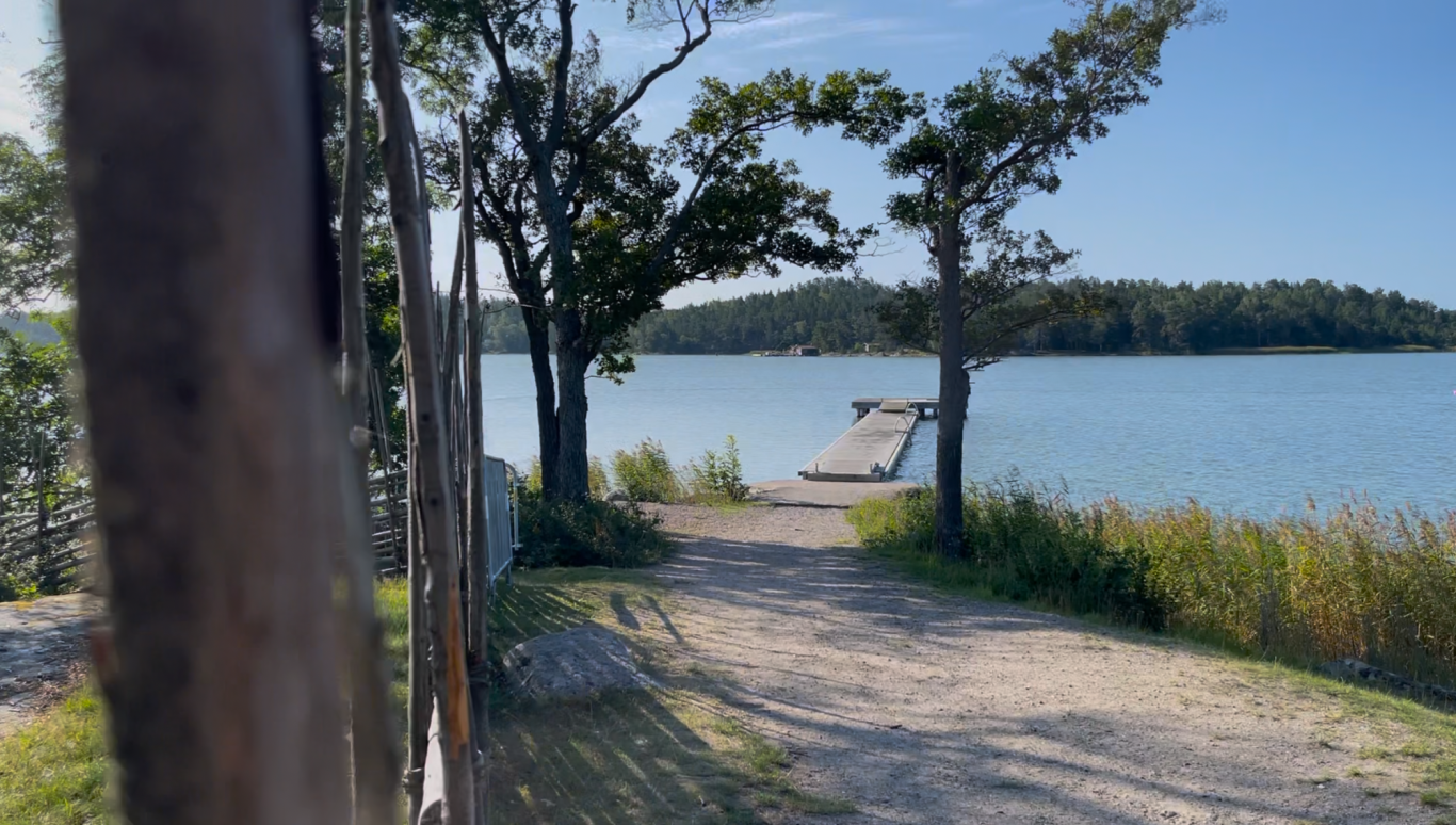 Badbryggan med Hopptorn En grusväg leder till en avlägsen brygga vid en sjö omgiven av träd och grönska.