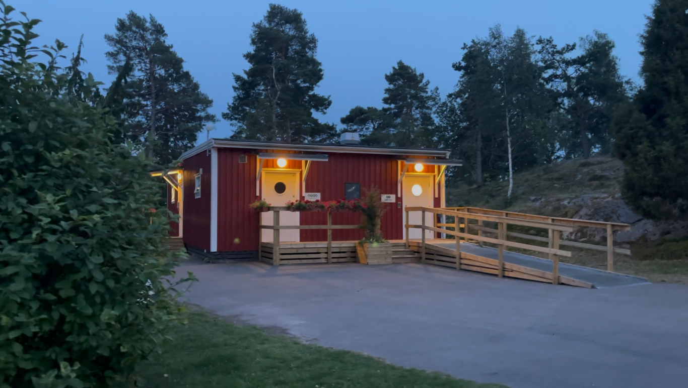 Toaletterna Röd stuga vid skymning med upplysta fönster och ramp, omgiven av träd.