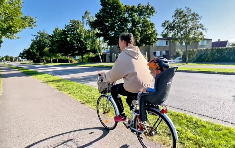 En cyklist med ett barn i en cykelsäte åker längs en grönskande gata.