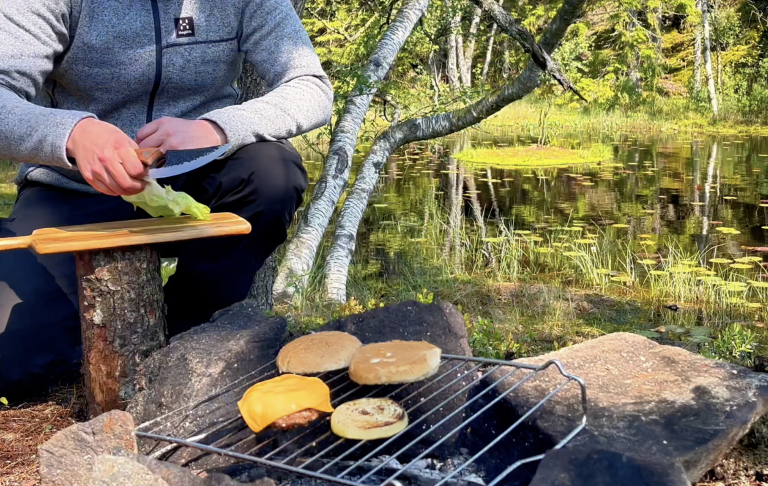 Person grillar bröd och ost vid en sjö omgiven av träd och grönska.