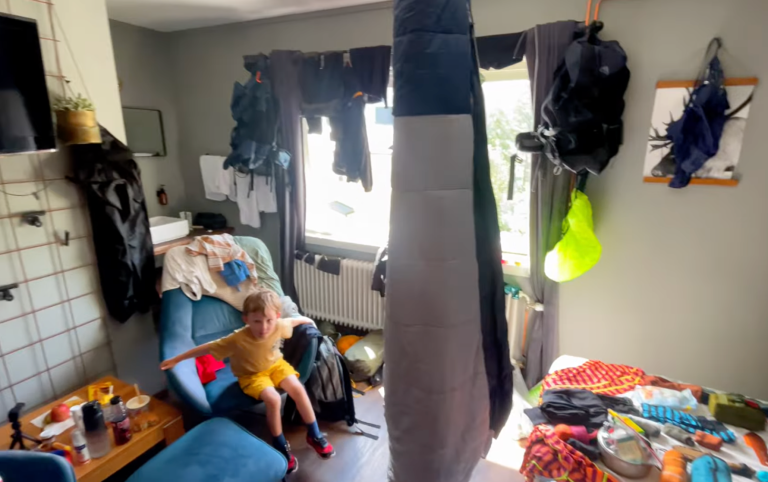 Rörigt rum med kläder och prylar utspridda, ett barn sitter på en soffa.