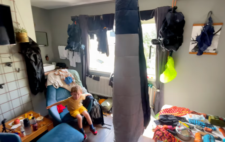 Rörigt rum med kläder utspridda och ett barn som leker på golvet.