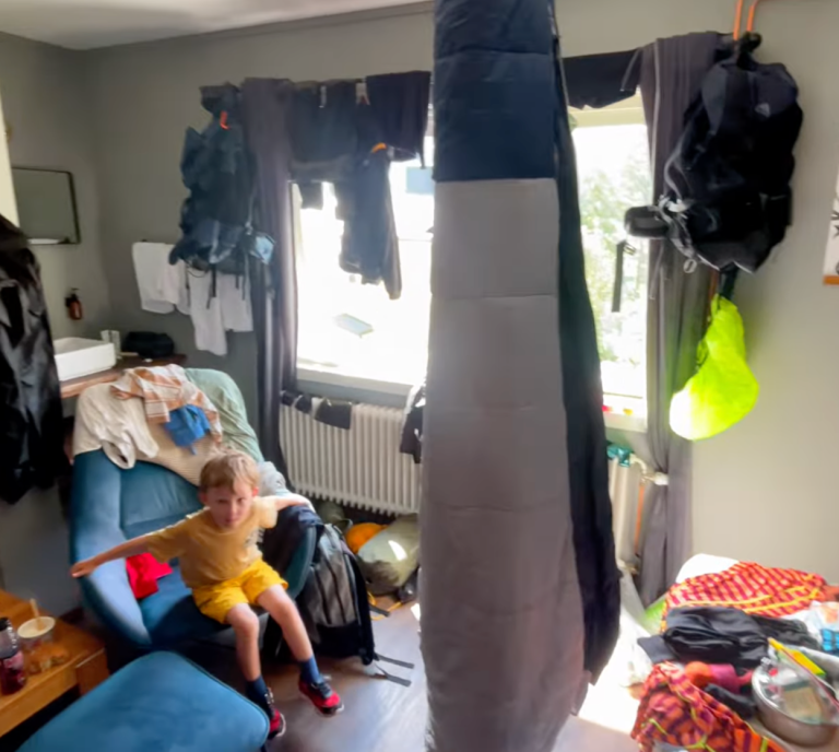 Ett barn sitter på en soffa i ett rörigt rum med kläder och föremål hängande omkring.