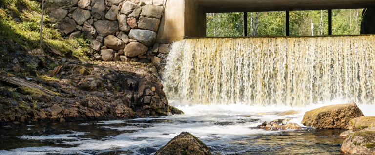 Ett vattenfall rinner under en betongbron, omgiven av stenar och grönska.