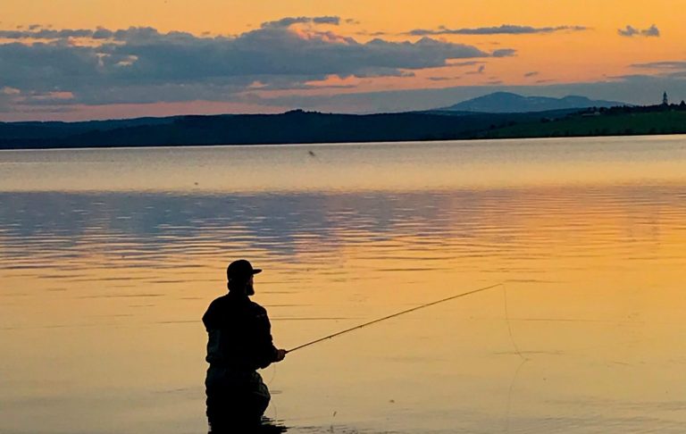 En siluett av en person som fiskar vid en sjö vid solnedgång med Åreskutans siluett i bakgrunden.