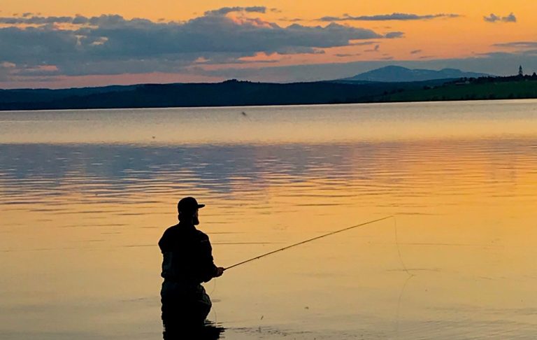 Fiskare står i vattnet vid solnedgång, med spö i handen i Jämtland med Åreskutan på horisonten.