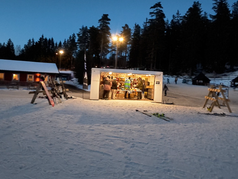 Uteplats i snön med människor och belysning i skymningen. Träd i bakgrunden.