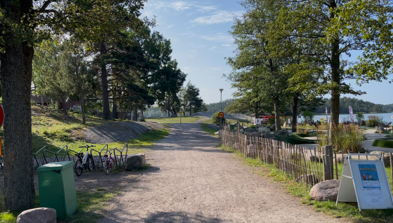 Algots på Udden och minigolfen Grusväg omgiven av träd, leder mot vatten och soligt landskap.