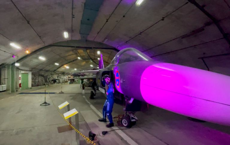 En tekniker står bredvid ett stridsflygplan i ett upplyst hangar.