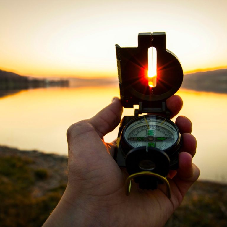 Hand holding a compass with sunset reflecting on water in the background.