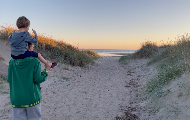 En vuxen bär ett barn på axlarna, som ser mot havet vid skymning.