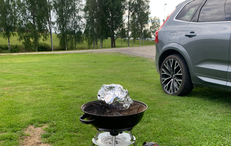 Grillar brevid bilen Grill med foliepaket på gräsmatta, parkering av en grå bil i bakgrunden.