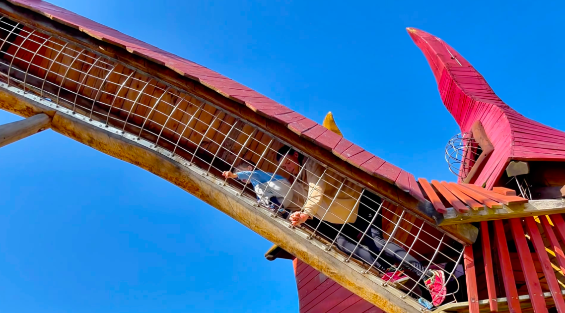 Vallarnas Friluftsområde Lekplatsen En röd rutschkana med en person som åker ner under en klarblå himmel.