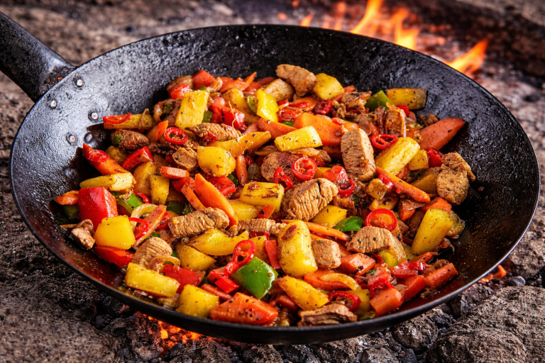 En stekpanna med stekt kött och grönsaker i olika färger över öppen eld.