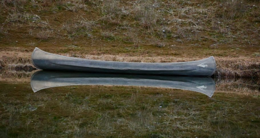 En kanot vid stranden En kanot vilar stilla vid vattnet, speglad i ytan, omgiven av grönskande land.