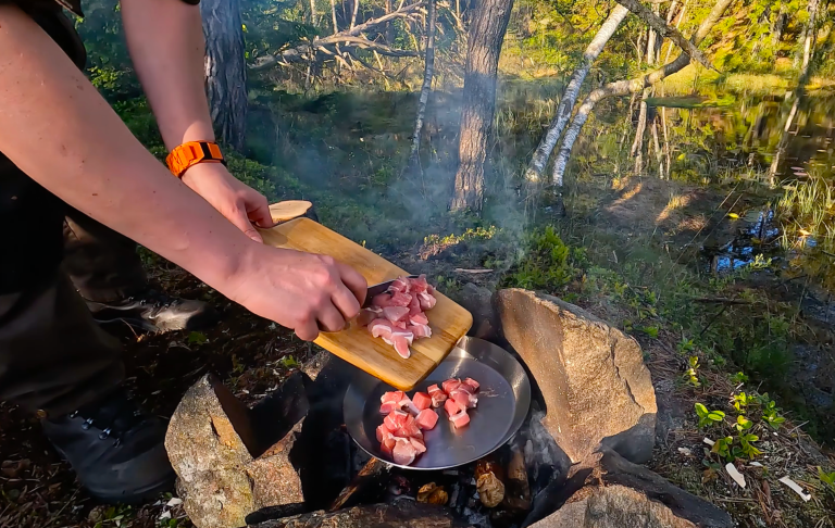 En person hackar kött på en skärbräda ovanför en eld i naturen.