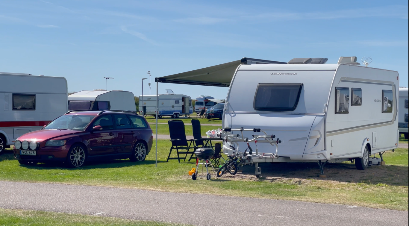 Björkängs Husvagn Camping En husvagn parkerad bredvid en bil, med stolar och ett markisad för solskydd.