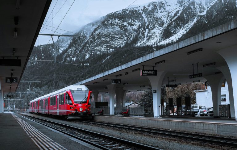 Röd tåg på en tågbana vid en modern station med fjäll i bakgrunden.