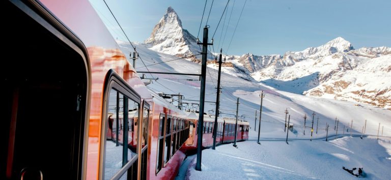 Tåg som passerar snötäckta berg med en spetsig bergstopp i bakgrunden.