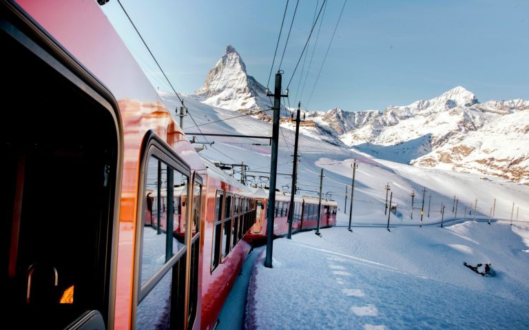 Röd tåg passerar snöklädda berg under en klarblå himmel.