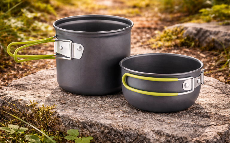 Två svarta campinggrytor i aluminium med gröna handtag, placerade på ett stenunderlag.
