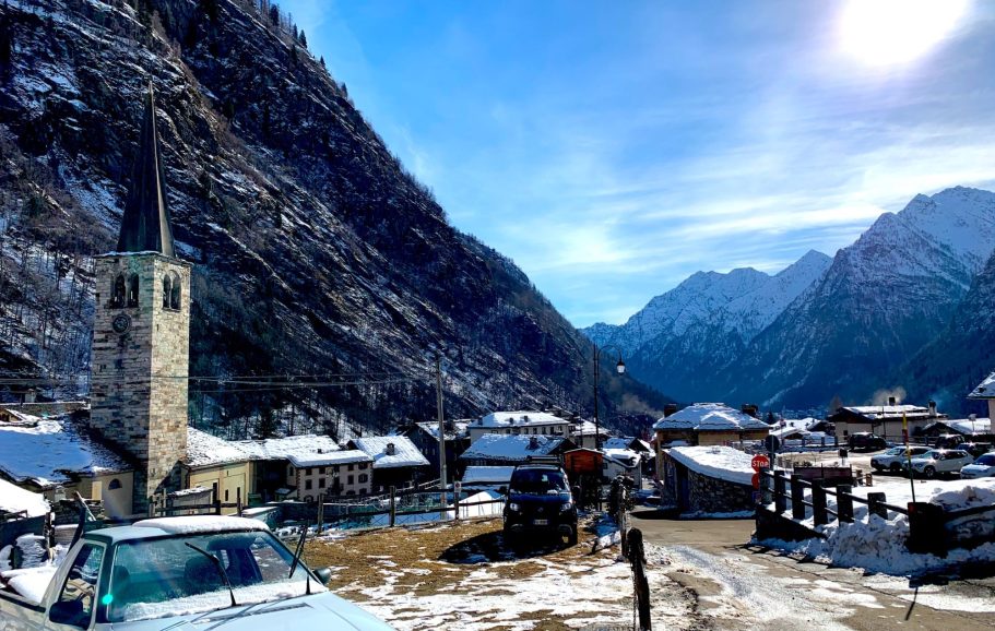 Snötäckta alpberg och en kyrka i en pittoresk Italiensk alpby under en klarblå himmel.