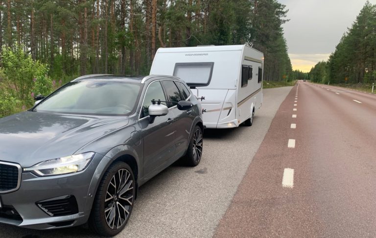 En grå SUV med husvagn parkerad vid en landsväg omgiven av träd.