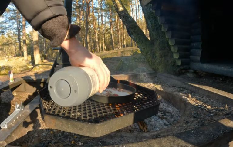En hand häller kolbullesmet ur en kanna över en grillad fisk vid en skog.