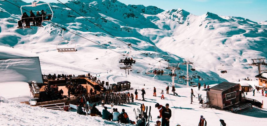 Folk åker skidor i en snötäckt skidbacke med liftar och fjäll i bakgrunden.