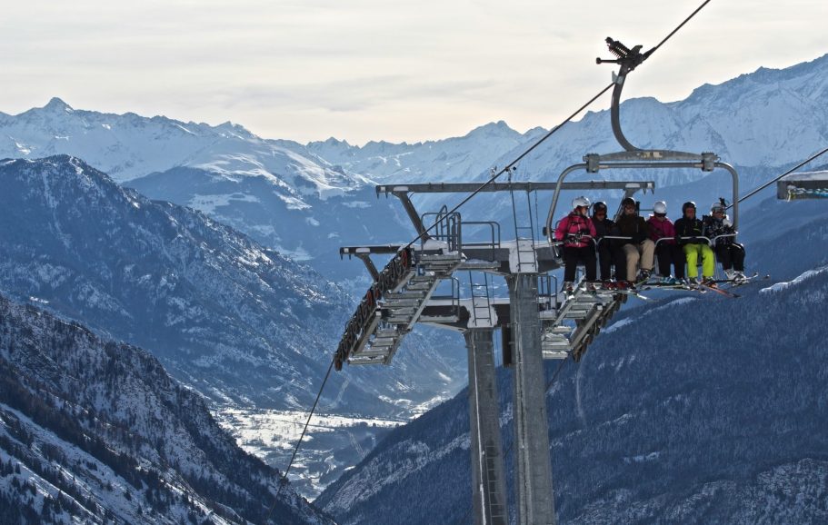 Skidåkare åker upp i en stolslift med snötäckta berg i bakgrunden.