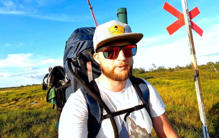 En man med ryggsäck och keps står på fjället längs en vandringsled med blå himmel i bakgrunden.