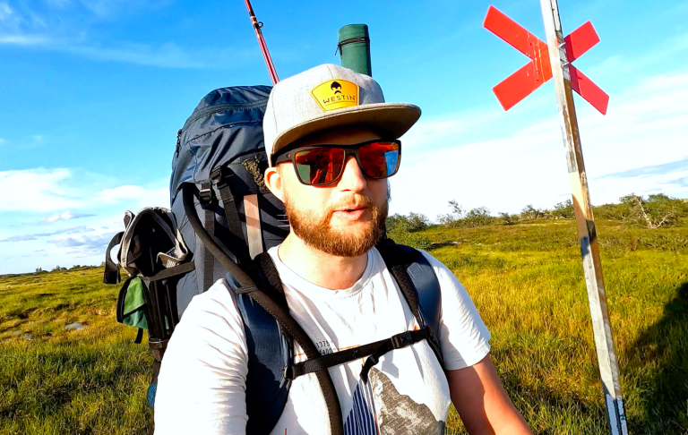 Man med ryggsäck och glasögon, vandrar på en grön landskap med en röd vägskylt.