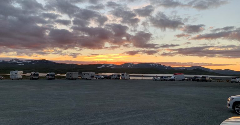 Solnedgång över fjällen med mörka moln och parkerade bilar i förgrunden. Den gamla gruvan i bakgrunden.