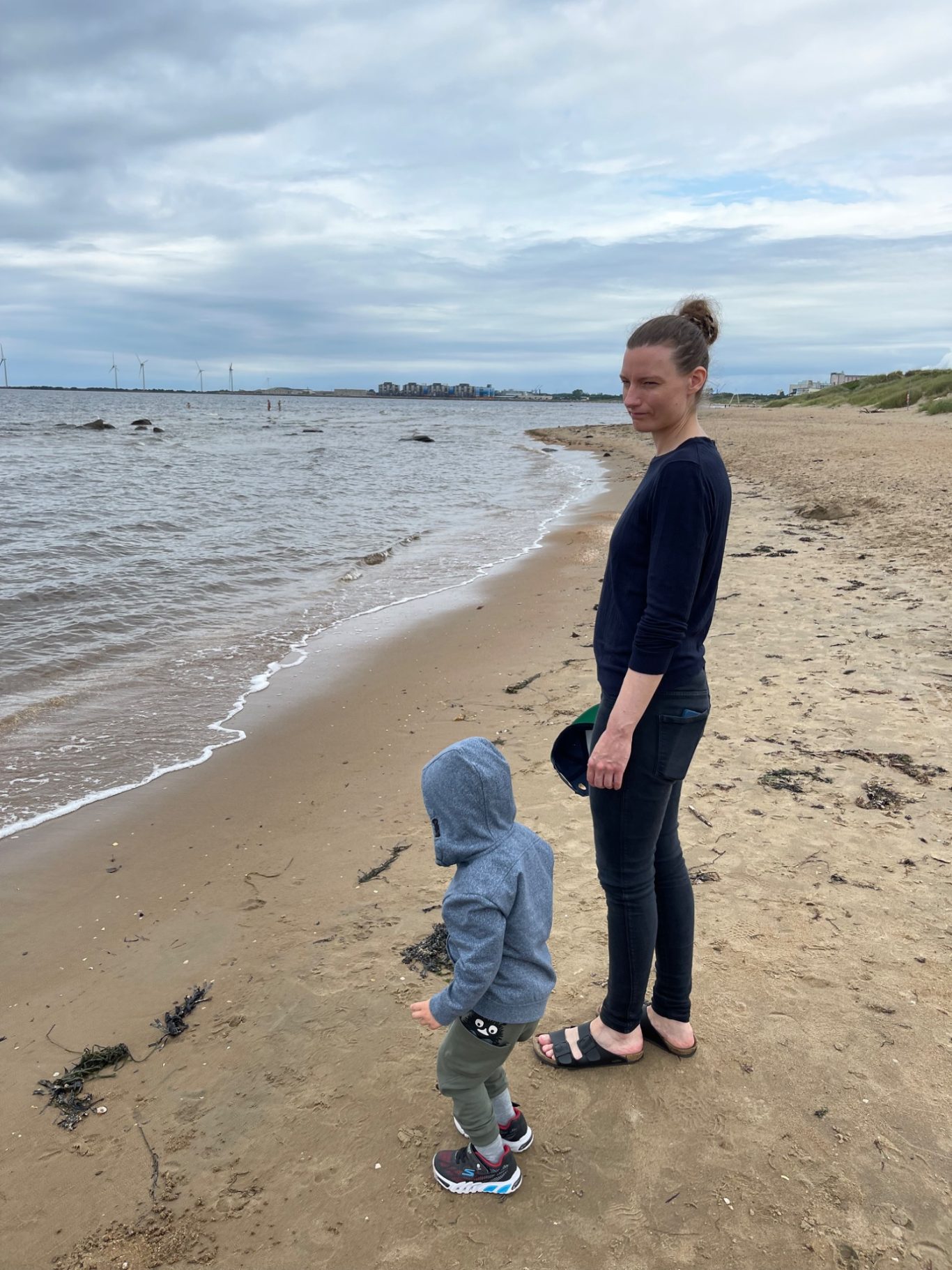 Skrea Strand Kvinna står vid stranden bredvid ett litet barn, båda tittar ut över vattnet.