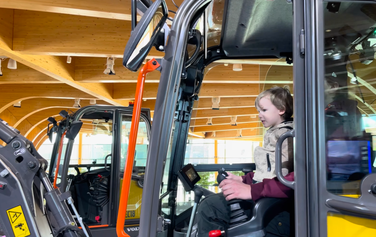 En person sitter i förarhytten av en grävmaskin inomhus.